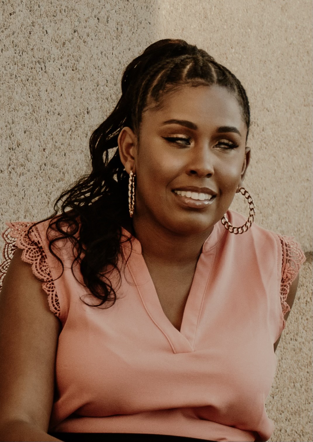 A woman smiling, wearing a pink blouse with lace sleeves and large hoop earrings. She has long, wavy hair tied back and is sitting against a textured wall.