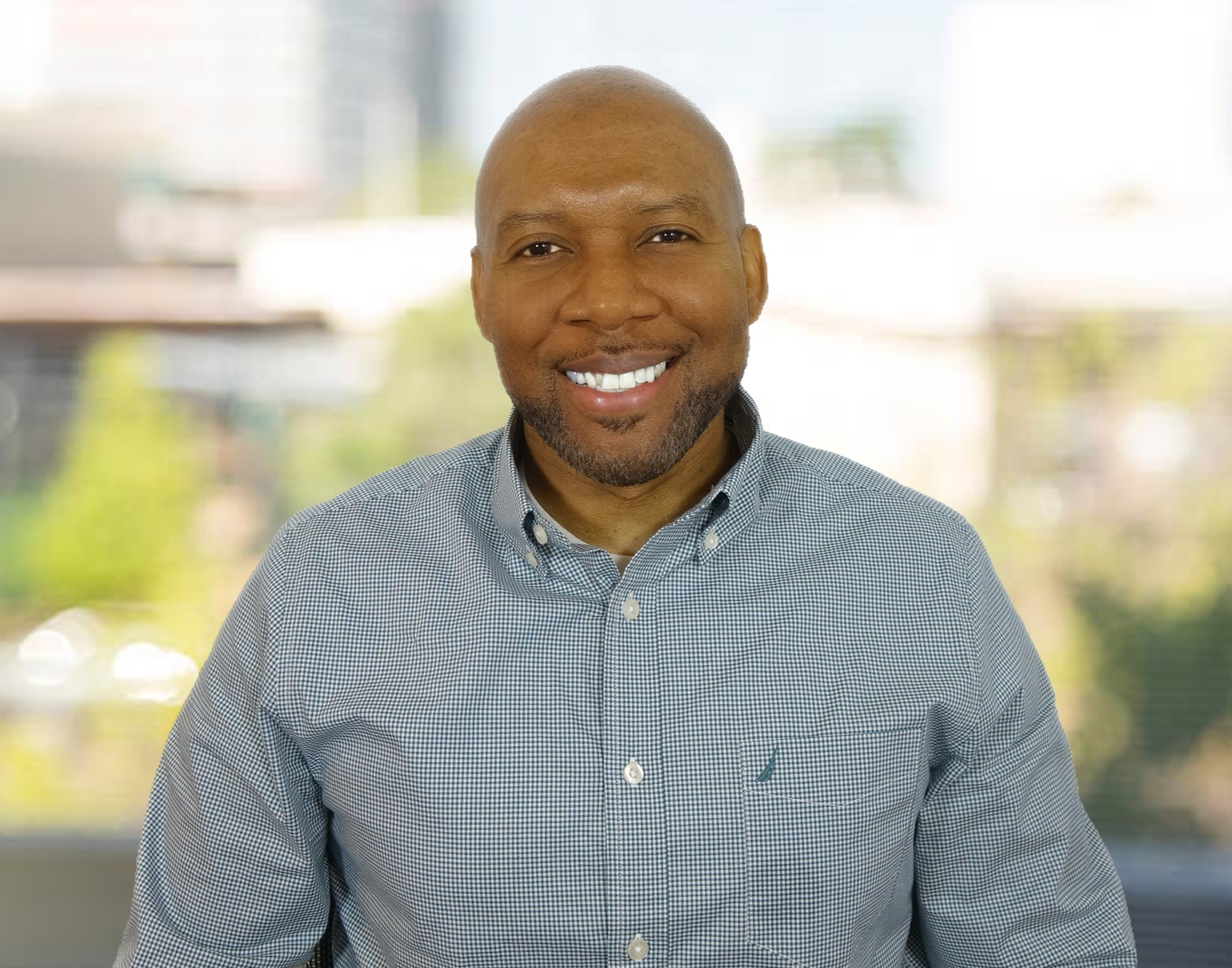 A person smiling at the camera, wearing a checkered button-up shirt. The background is blurred, showing outdoor scenery with buildings and greenery.