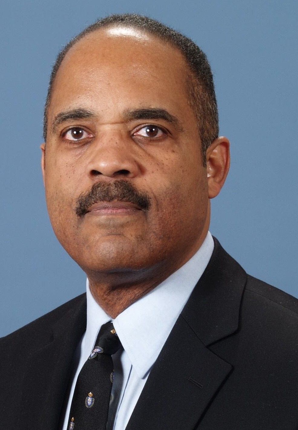 A man in a formal black suit, white shirt, and patterned tie is looking directly at the camera against a plain blue background. He has a mustache and short hair.