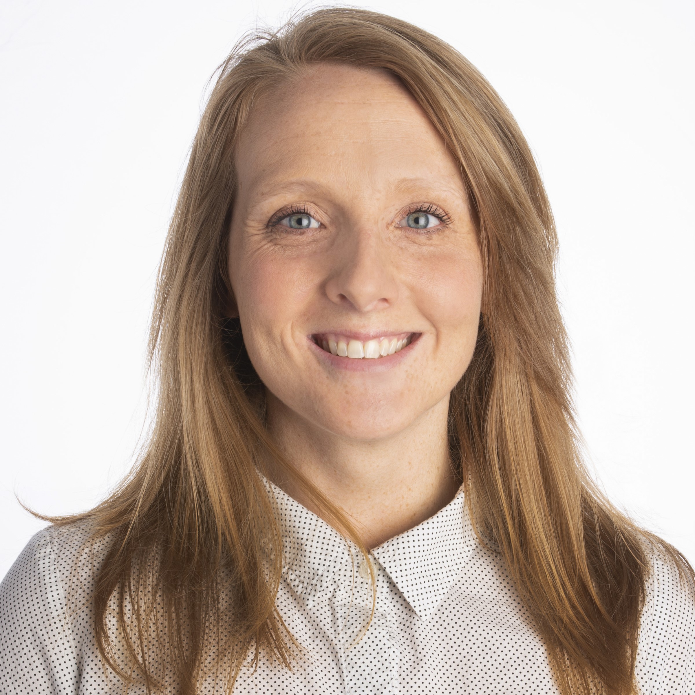 A person with long, straight hair is smiling, wearing a light-colored, patterned shirt. The background is plain white.
