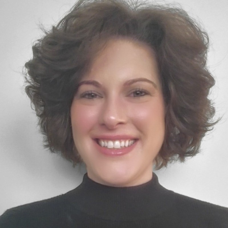 A person with short, curly brown hair is smiling at the camera. They are wearing a black turtleneck top and are set against a plain white background.